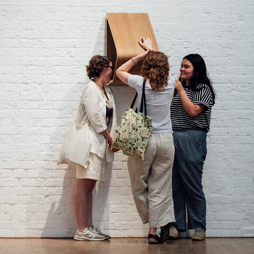 Three adult females listening closely to a wooden wall-mounted sculpture