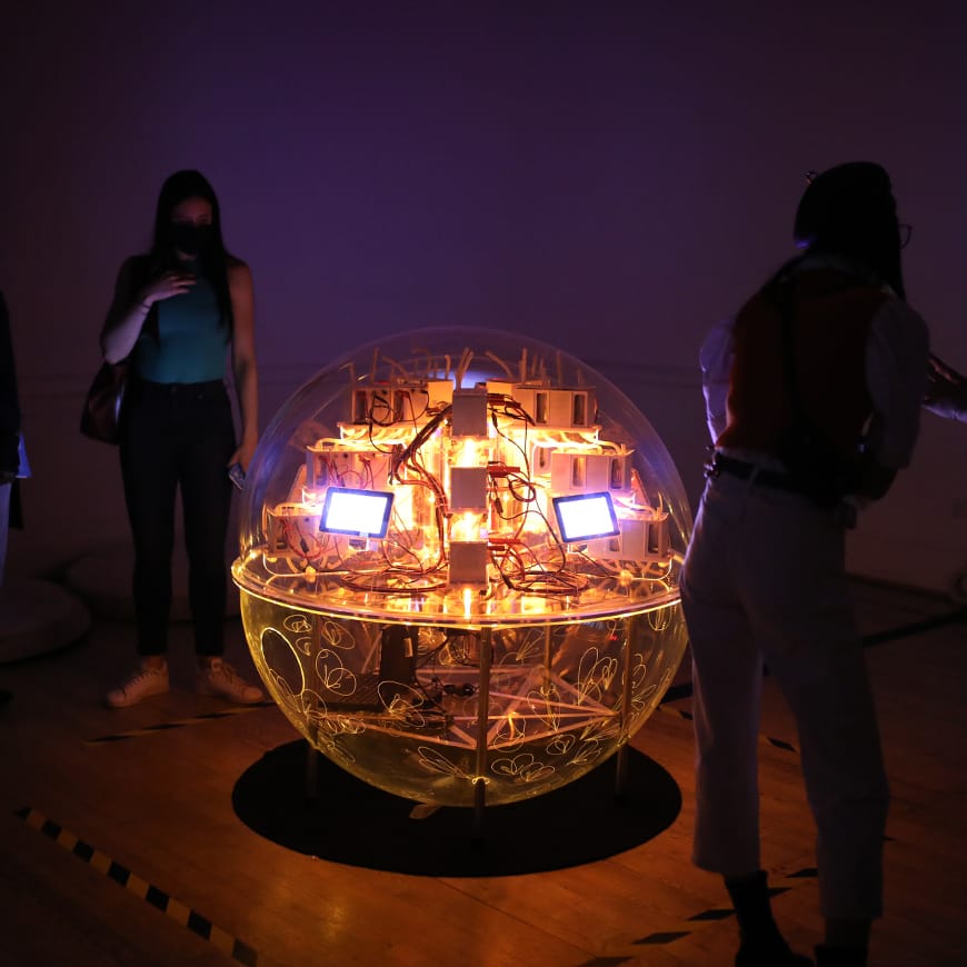 Two people in a darkened room by a transparent sphere containing electronics and LEDs and lit by an internal amber glow