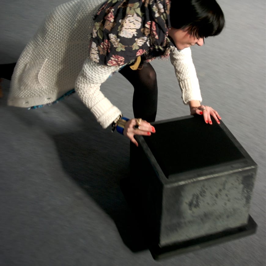 A person with dark hair and red nails pushes the black cube across the gallery floor
