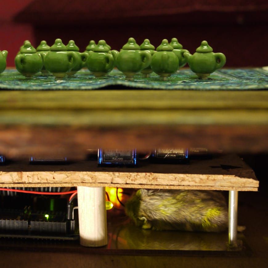 Side view of tiny green teapots sitting in top of electronics board and taxidermy hamster